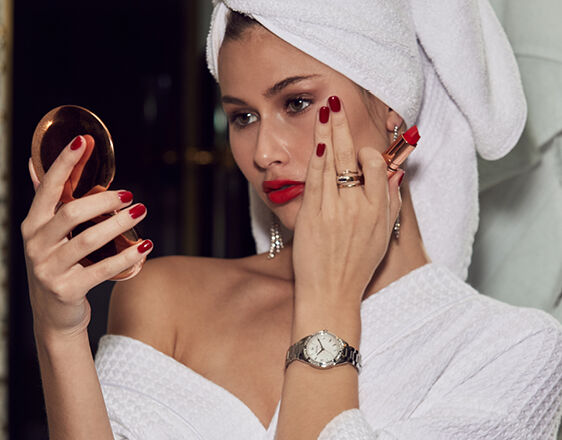 Elegant woman in towel applying lipstick while wearing Bulova ladies’ stainless steel watch with white dial.